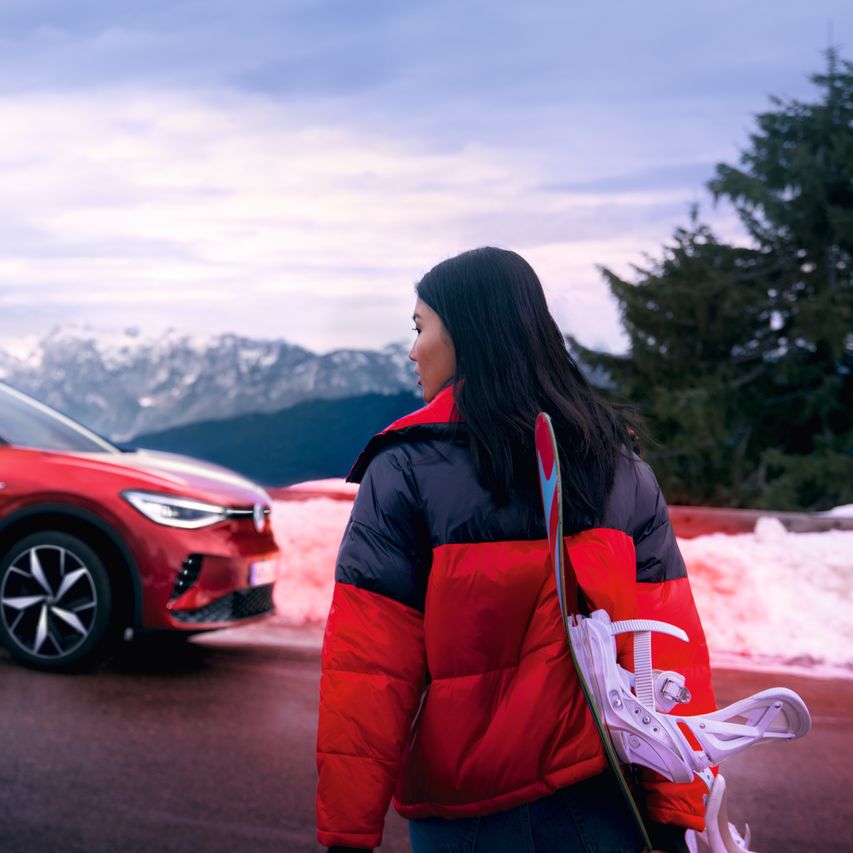 Страничен изглед на червен VW ID.4 GTX на планински фон, на преден план жена гледа към автомобила