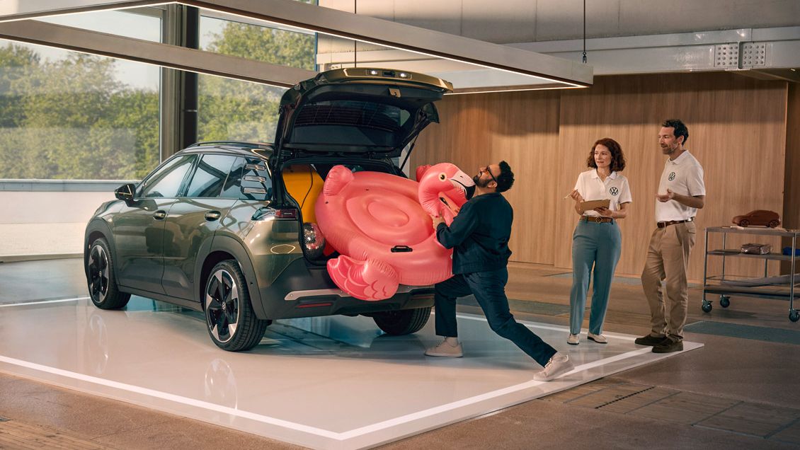Мъж бута надуваемо фламинго в отворения багажник на VW ID. Кръст. Двама души стоят до него и го наблюдават с усмивка.