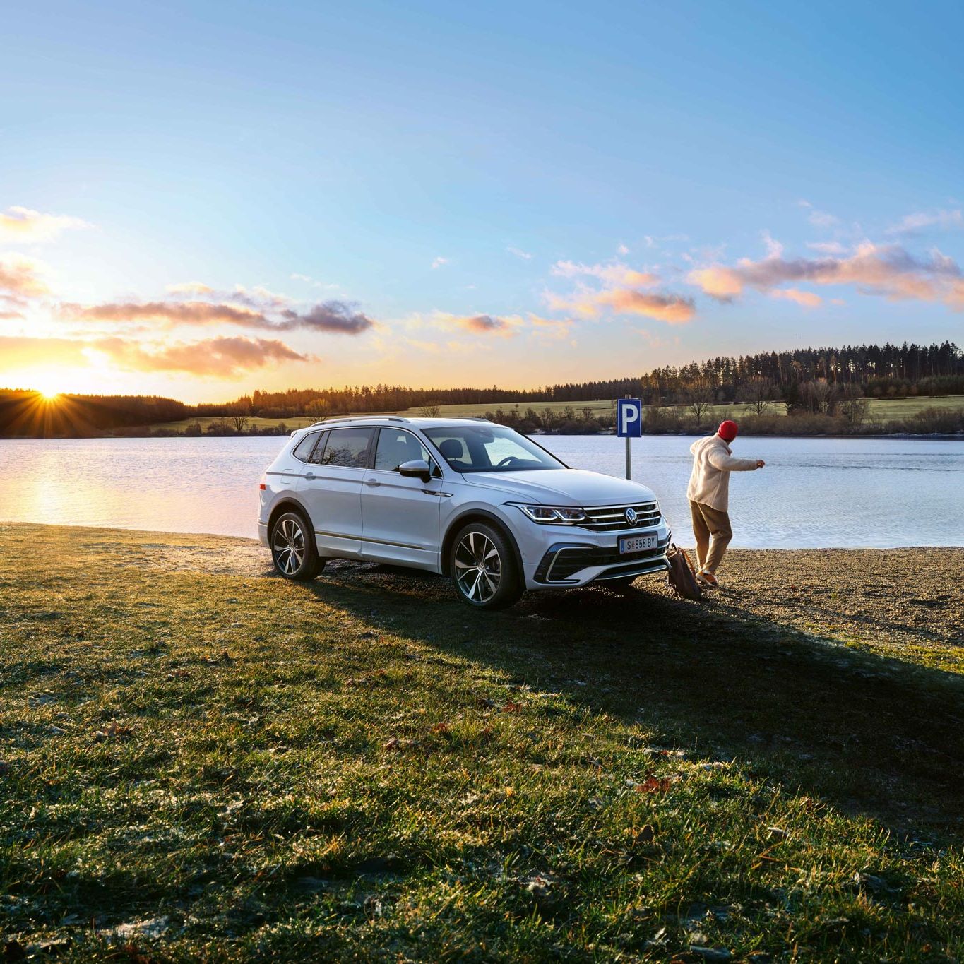 Бял Volkswagen Tiguan паркиран край езеро по залез с човек, който се разхожда наблизо.