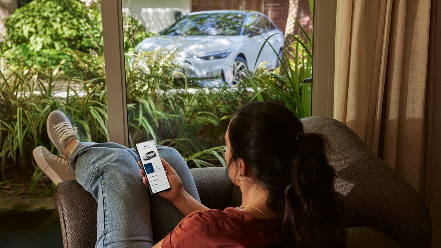 Eine Frau lehnt in einem Stuh und verwendet die VW App auf ihrem Handy, draußen ist ein weißer VW ID.7 geparkt