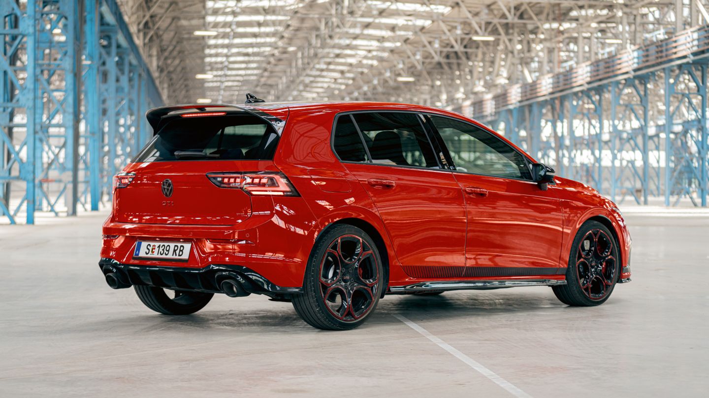 Ein roter VW Golf GTI steht in einer großen, lichtdurchfluteten Industriehalle. Das Auto ist von hinten und leicht seitlich zu sehen. Die schwarzen Räder und der Heckspoiler fallen deutlich ins Auge.