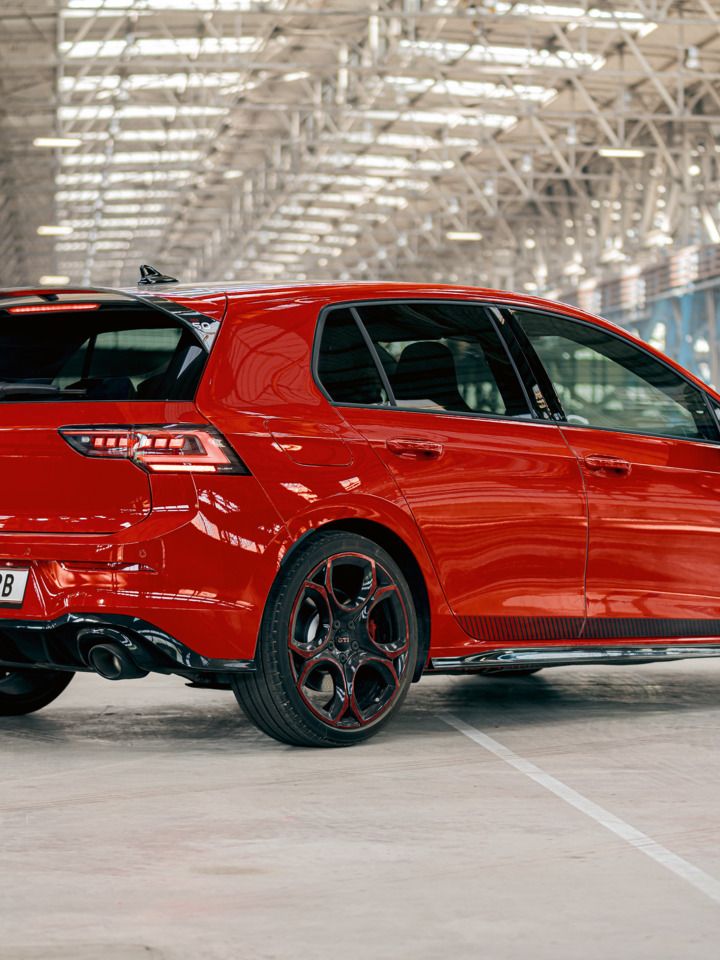 Ein roter VW Golf GTI steht in einer großen, lichtdurchfluteten Industriehalle. Das Auto ist von hinten und leicht seitlich zu sehen. Die schwarzen Räder und der Heckspoiler fallen deutlich ins Auge.